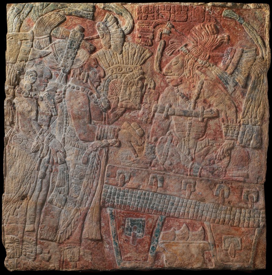 A carved stone panel showing a seated figure in a huge feathered headdress on a throne, other figures facing him, and blocks of small square symbols above.